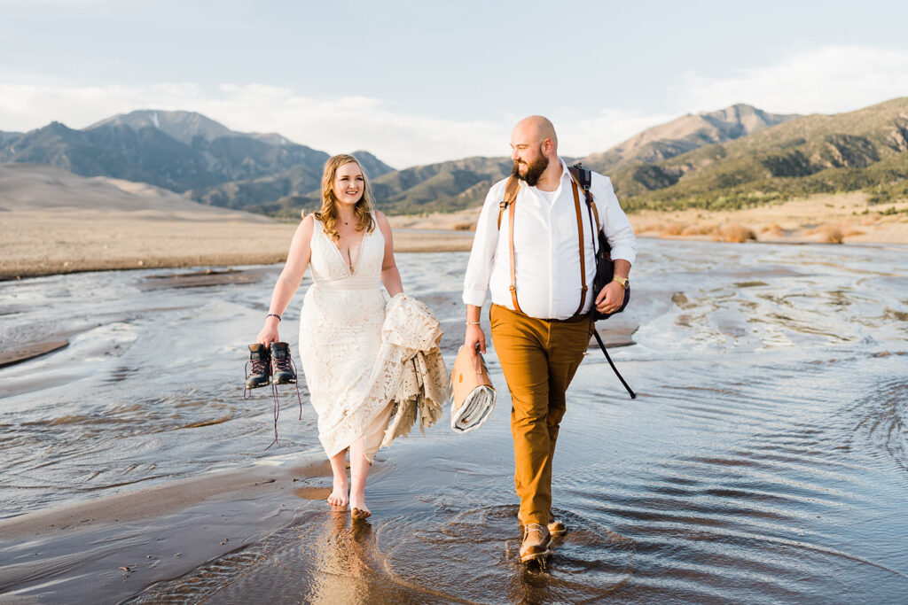 bride and groom walking with bare feet through river in front of the Great Sand Dunes during their adventure elopement in Colorado