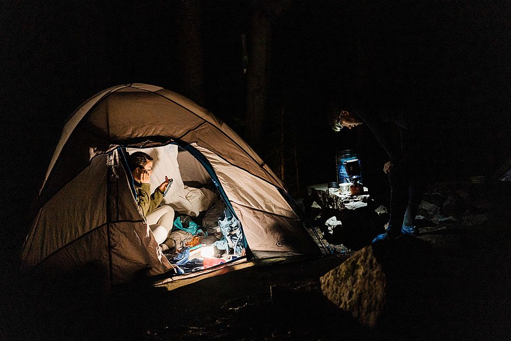 bride sitting in tent in the dark lit up by a flashlight doing her makeup while the groom is wearing a headlamp and making coffee in a jetboil outside the tent during adventure elopement in Colorado